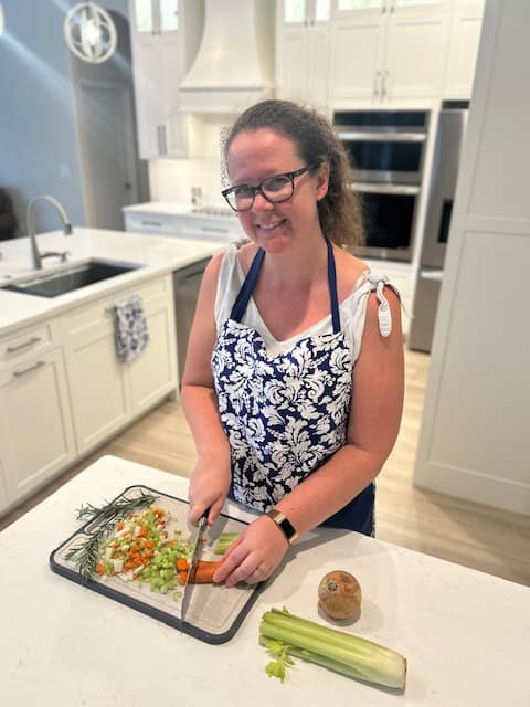 Char from Postpartum by Char lovingly chopping up vegetables such as carrots and celery to go into a pot of yummy homemade chicken soup. She is smiling in a pristine kitchen and wearing a blue floral apron.